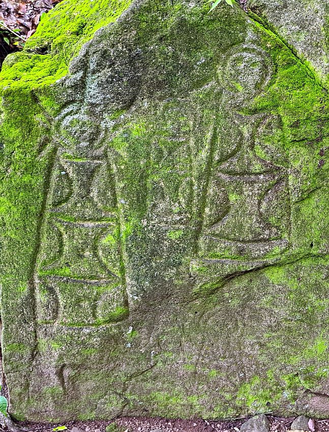 A flat face of rock with two figures: each as a round head at the top from two concentric circles. The bodies are more or less rectangular, with some simple designs carved in them. They're covered iwth a layer of moss. Some fainter carvings are around them, barely visible. It looks like there were smaller similar figures.