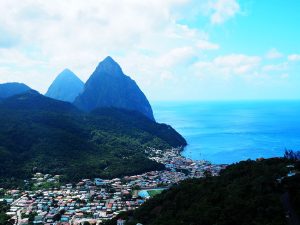 A view from a hill looks across a valley. In the valley a lot of houses, quite close together, in bright colors. They extend up the valley and also down right to the sea on the right. The sea is deep blue. A forest-covered hill rises on the other side of the valley and, beyond that, two taller pointed hills, one partly behind the other: the Pitons in Pitons Management Area.