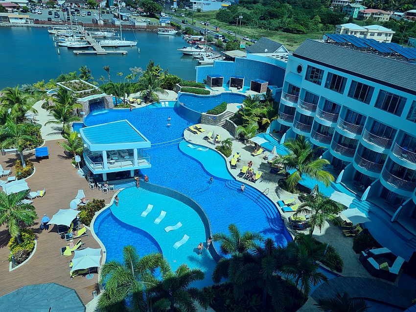 A freeform shaped pool with blue water, seen from above. Some sections are lighter blue because more shallow, with white loungers standing in the shallow water. A small two-story structure on pillars holds the bar. Lounge chairs and palm trees hear and there. Marina in the background and part of the Harbor Club hotel visible on the right, 4 stories of rooms with balconies.