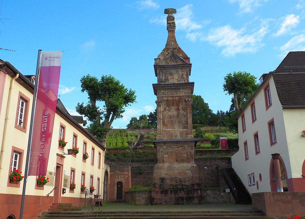 The column stands in a small open plaza between two 3-story buildins and is nearly twice as tall as they are. It is square and carved with a variety of images.