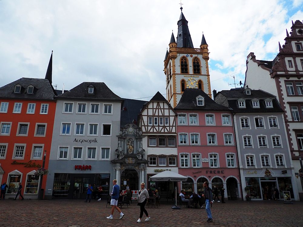 On a plaza in Trier, a row of pretty buildings, each painted a different color and of varying height. Behind them a tower is visible of a church. An ornate little entrance to the church is between two of the buildings. 