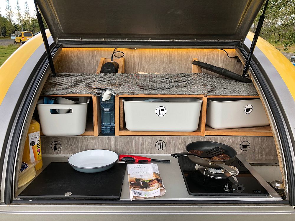 The open back of a teardrop shaped camper. It shows storage bins labeled for cutlery, pots, etc. In front of them, a flat space with one burner and work space.