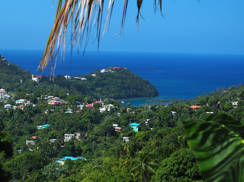 A view from a hill on Saint Lucia looking over a very green and hilly landscape with a scattering of houses painted in pastel colors. In the distance, the deep blue sea.