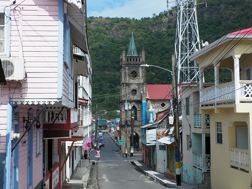 Looking down a road in Soufriere, Saint Lucia. houses of various heights in pastels line the sides. A stone church tower further down the road. The pavement is not wide and neither are the sidewalks and both are patched.