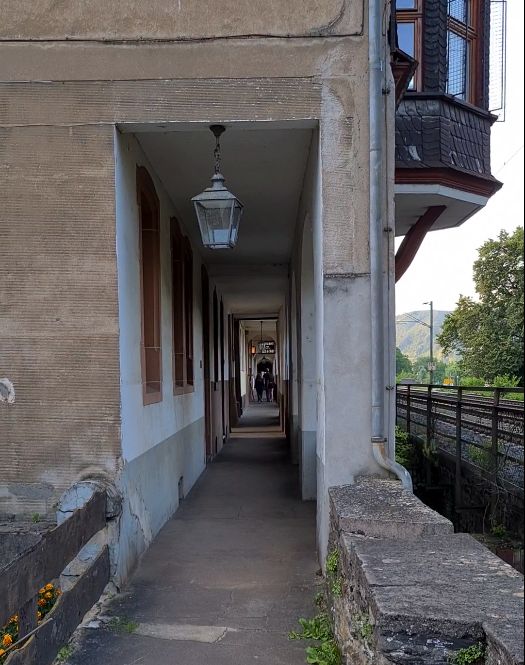 Looking down a narrow walkway, wide enough for two people to walk next to each other if they stay close together. Wall on the left has windows in it. Roof overhead, then square simple columns on the right. The bottom of a bay window is visible sticking out beyond the colums, and a number of railroad tracks parallel the walkway.