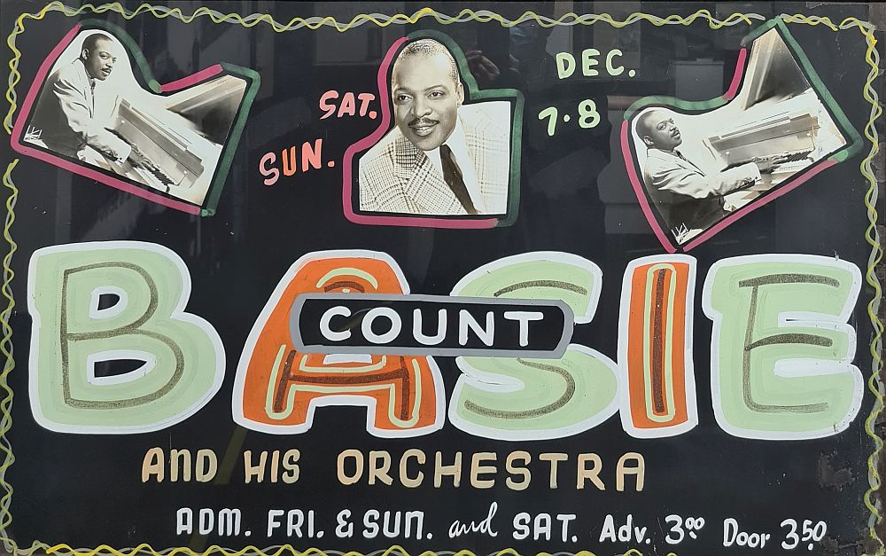 An ad for a performance by Count Basie and his orchestra. It looks like it would be the kind that's hung right in front of the venue and is written in what looks like paint or chalk, with a few photos of Count Basie tacked onto it. 