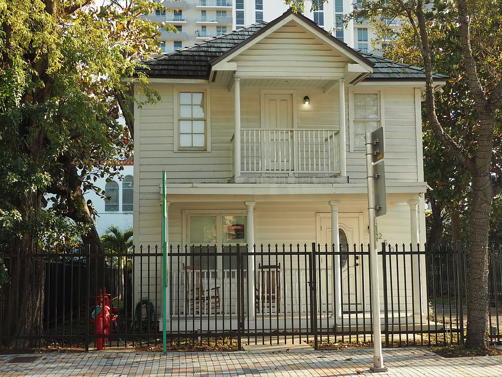 A simple, white painted wooden house, two stories, with a shaded front porch with 2 rocking chairs. Upstairs has a smaller porch. The house is sided with horizontal panels and has a tree on either side, and a much taller apartment building behind it.