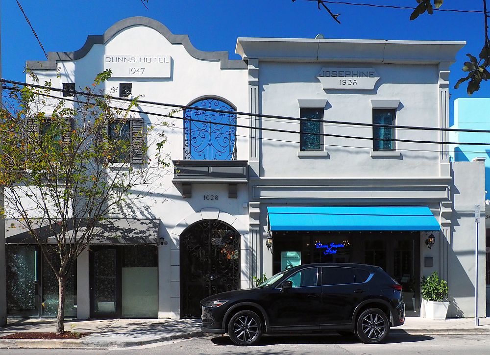 Both buildings are 2 stories. The one on the left has a plaque at the top reading "DUNNS HOTEL 1947" and the right-hand one says "JOSEPHINE 1938". Both are painted in neutral shades of white and slightly gray. The one on the right, the entrance to the Dunns-Josephine Hotel, has a blue awning over the sidewalk and a blue neon sign in the window that names the hotel.