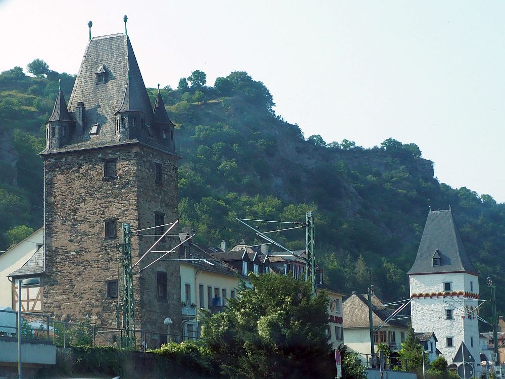 On the left, a tall square tower with a pointed roof. On the right, more distant, another tower shaped the same, with a similar roof. The near one is stone, the far one is probably also stone, but it is painted or plastered white.