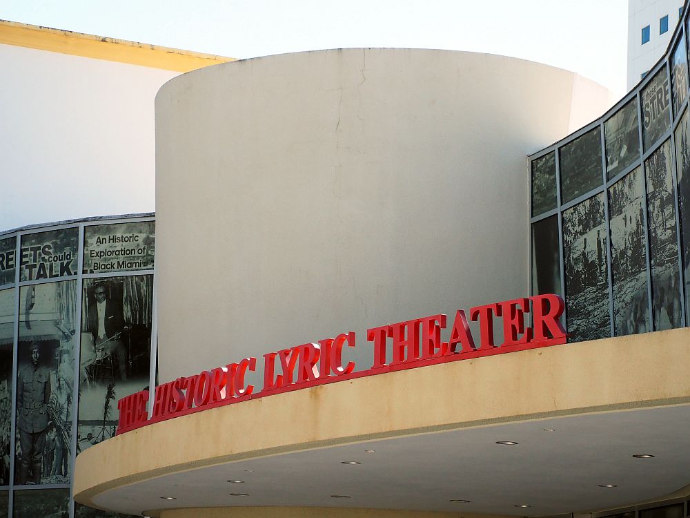 A white cylindrical shape, with the roof above the entrance below it. Along the roof are the words "The Historic Lyric Theater." On the wall on either side are many black and white photos of historical events in Historic Overtown.