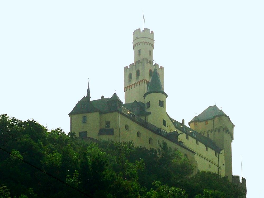 On top of a hill, this Rhine Valley castle looks white. It has a tall square tower with crenellations around its top, and on top of that square tower is an even higher cylindrical tower, topped by a flag. At the other end of the castle is another tower, rectangular this time, with a somewhat more modern roof.