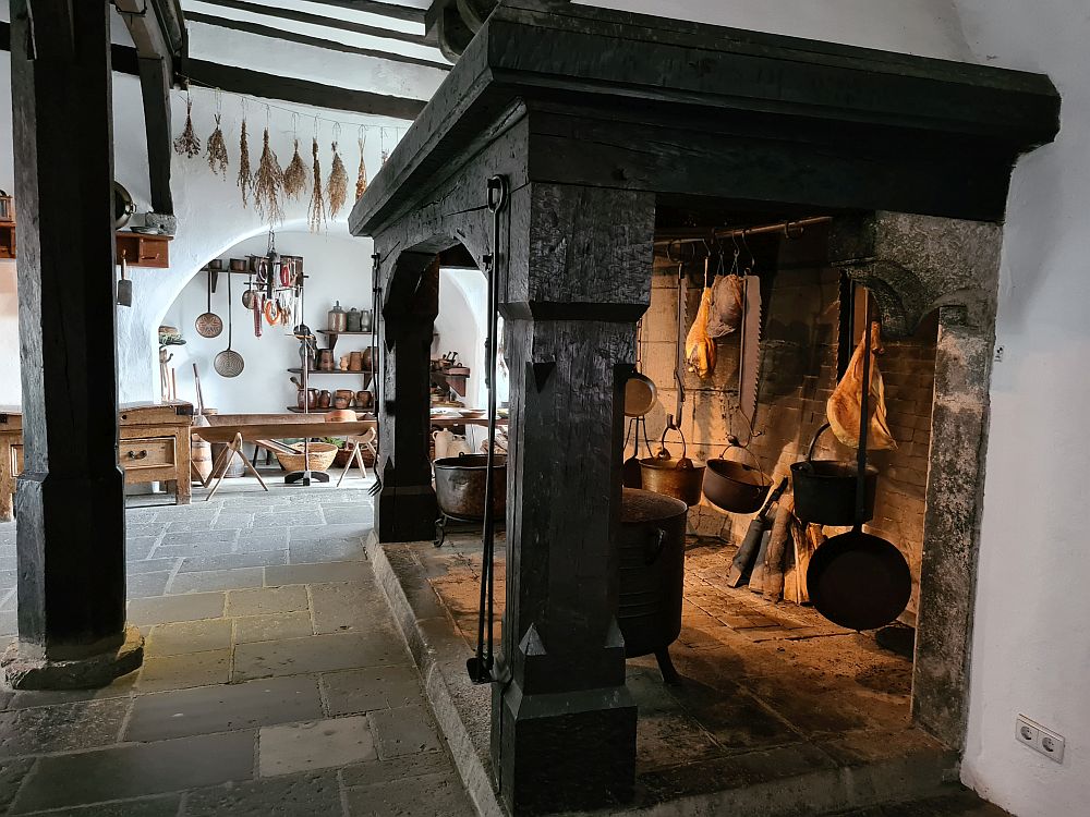 A huge hearth, with various pots hanging over it and hunks of meat as well (presumably fake). Beyond is a work area with lots of pots, pans, pitchers and other kitchen tools, with dried herbs hanging from the ceiling above.