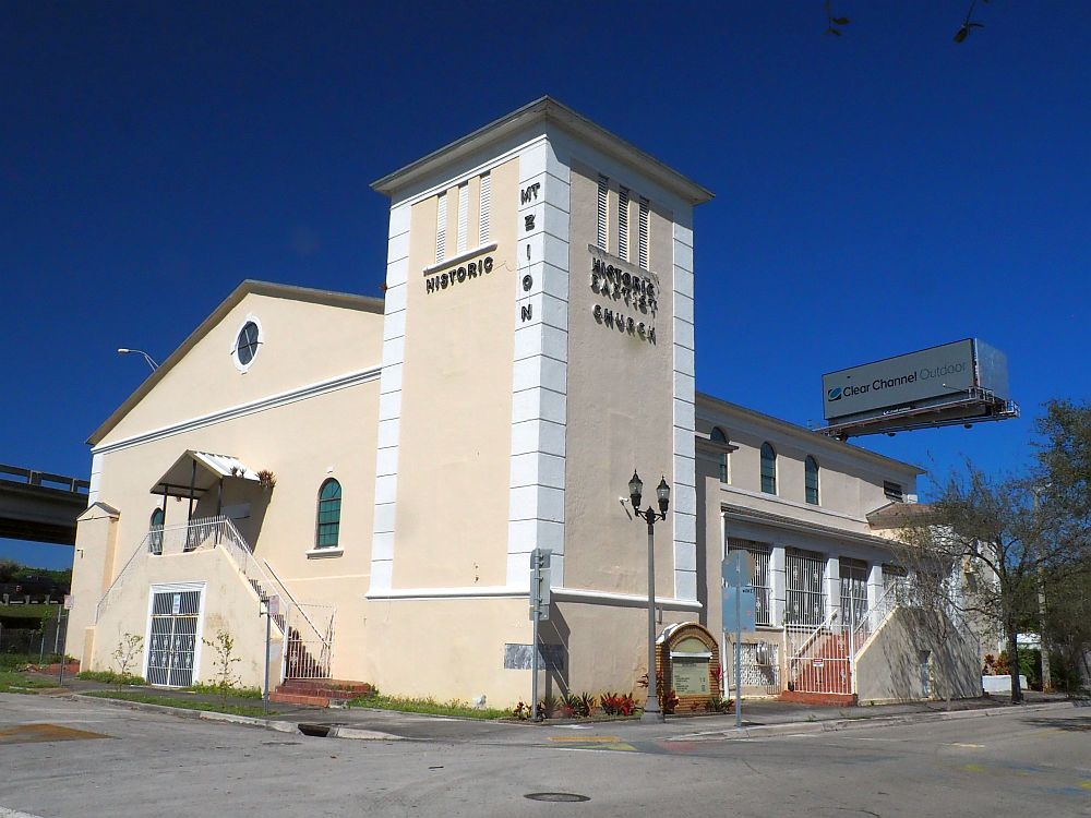 The church has a peaked roof and a square tower in one corner, not much higher than the roof. The church is painted beige with white trim. The highway is visible passing behind it and a huge electronic billboard as well. 