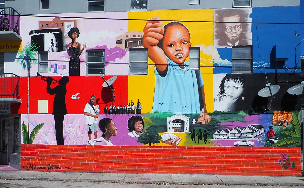 Painted in bright primary colors, and including several different images. In the center, a young boy holds out his fist. A female performer (I think, is in the upper left. Bottom left a man in silhouette plays a trumpet. A row of men whole rifles. An image of books, and also of the old Lyric Theatre and a row of shotgun houses too.