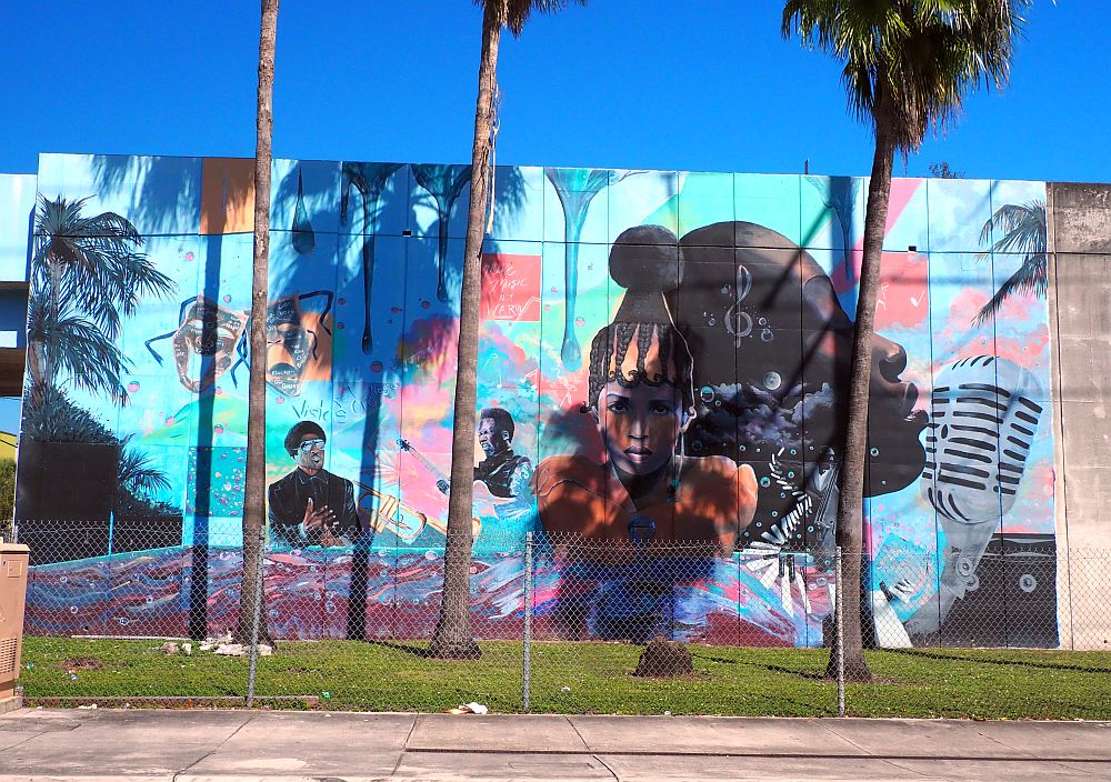 In the mural a number of musicians are shown as separate images, i.e. a woman singing into an old-fashioned microphone is depicted much larger than the others. A pair of theatrical masks are also visible. Three palm trees rise in front of the mural.