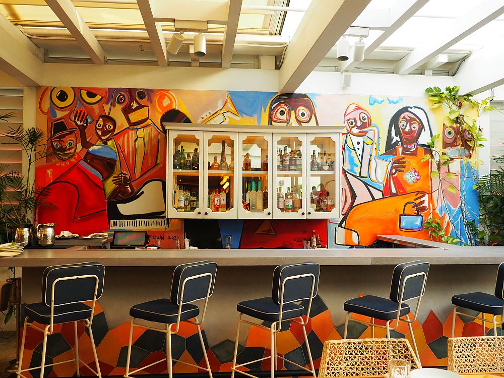 A bar inside the Red Rooster has a mural painted against its back wall, around a case holding the liquor. The images show musicians with instruments, in an almost cubist style.