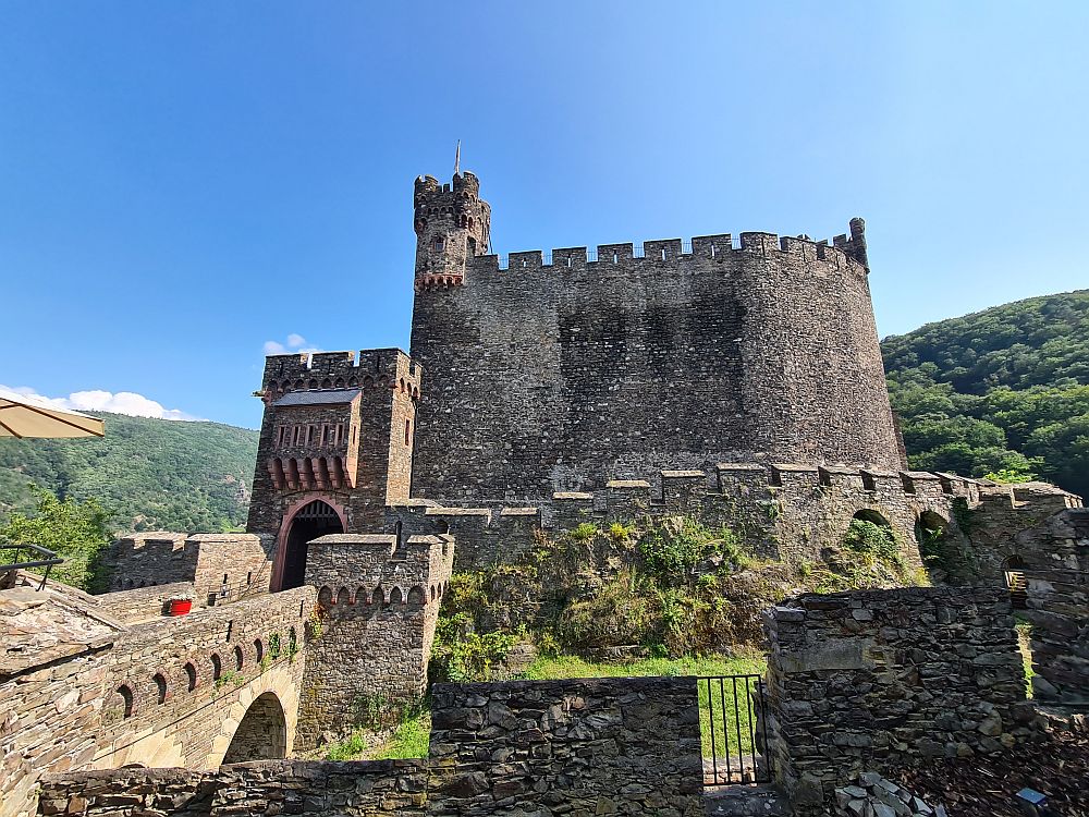 A stone wall, curved to an oval shape, very tall, with crenellations along its top and a small tower at one end. Another wall with crenellations at its food. A gate to the right, with an archway at the end of a bridge that arches over a dry moat.