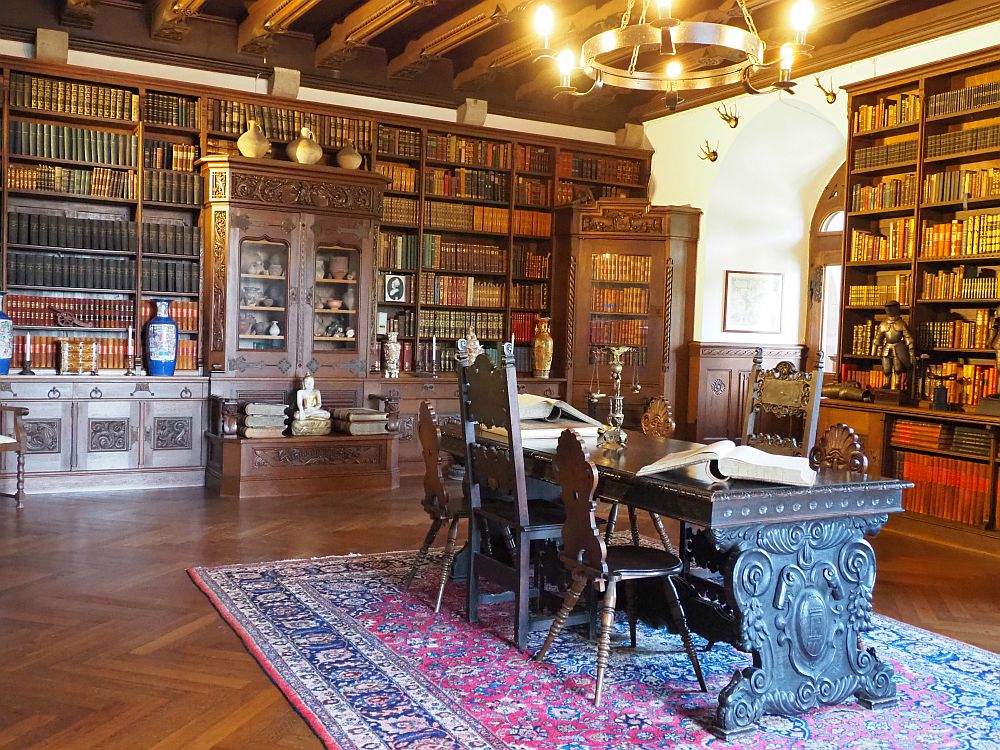 The entire far wall and what is visible of the right-hand wall are filled with dark shelves which are filled with books. A cabinet in the center of the far wall, built into the bookshelf, shows other objects through its glass front. In the center of the room, an oriental carpet, and on that, a heavily carved wooden table with six chairs around it.