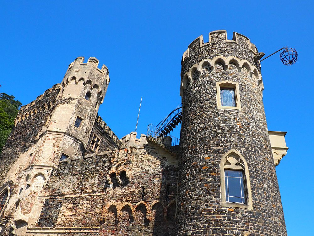 Rheinstein Castle, made of stone, with round towers with crenellations at the top. A metal stairway is visible leading up to the nearest tower, and extending out of this same tower is an odd metal structure: a rod sticking straight out and hanging from it, a metal basket of some sort.