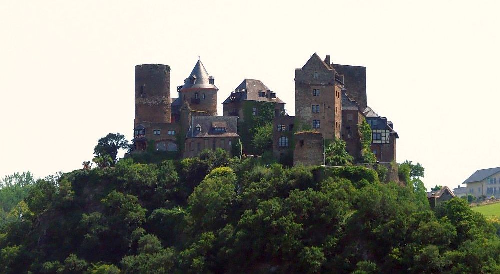 This Rhine Valley castle has lots of different structures making up the whole, many of which are visible, all grouped together on a hill, in this photo. A cylindrical tower on the left, a round tower with a pointed roof next to that, a rectangular block in the center, more rectangular blocks on the right, one of which has a half-timbered facade.