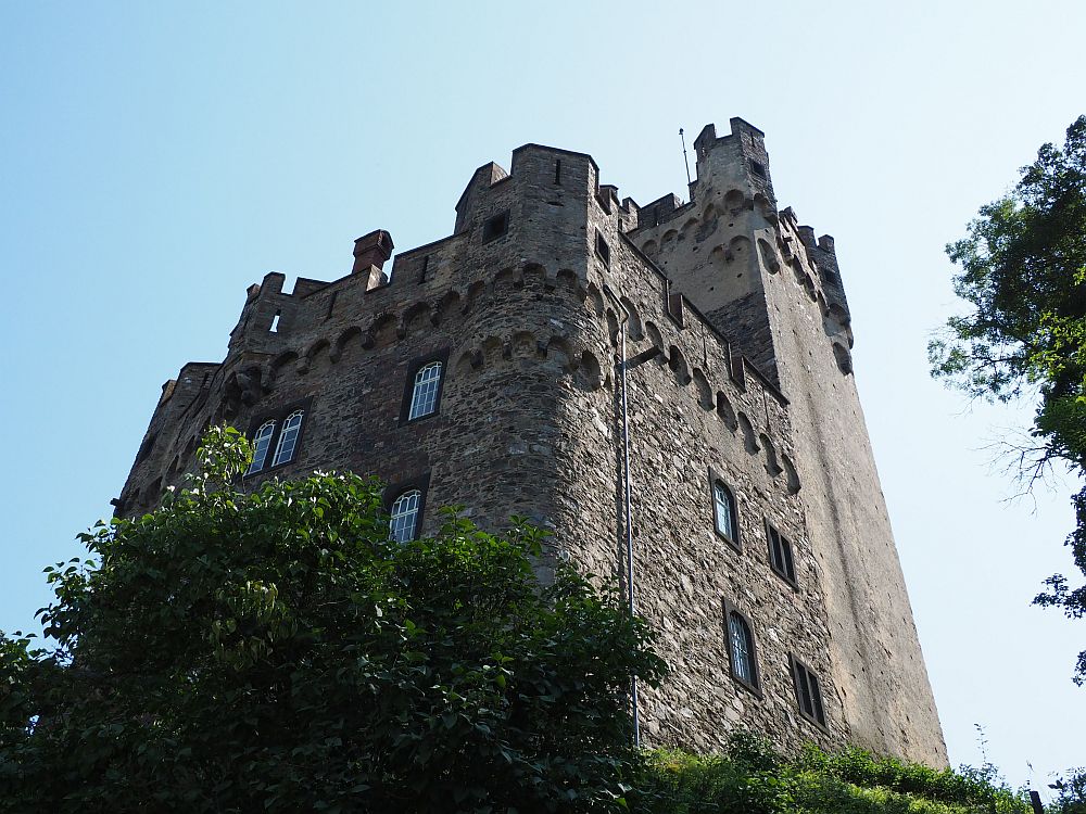 Looking upwards at a looming stone castle with crenellations at the top. The nearer part is rougher stone but looks newer because it has more windows. The rear part is taller, with towers on top, and has no windows.