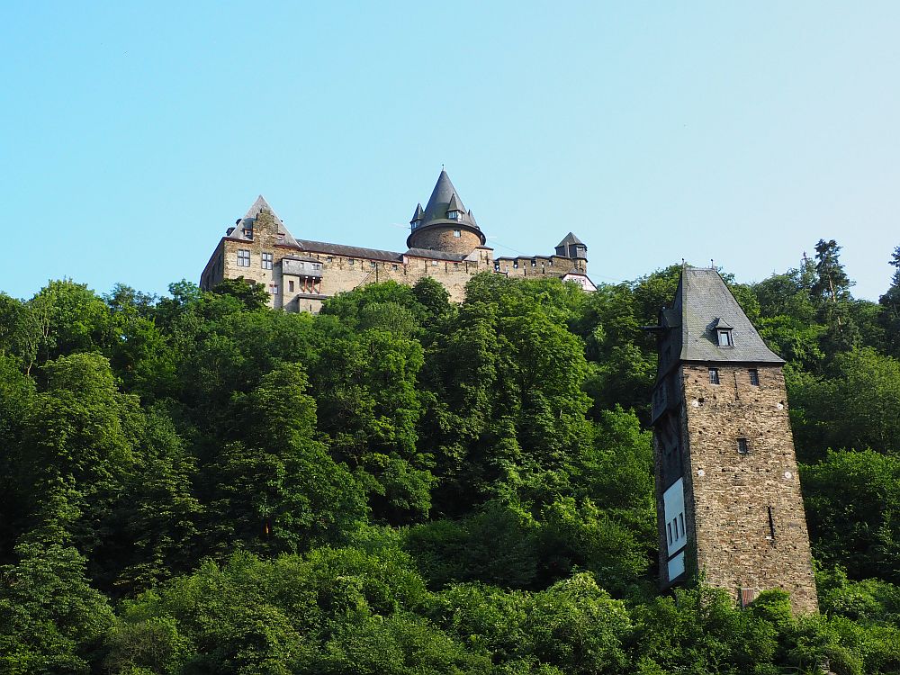 A wooded hill. On the right, a square, stone tower with a pointed roof. Above, on top of the hill, part of a castle is visible: a flat wall with crenellations, a few towers with round conical roofs.