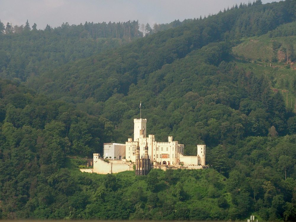 This Rhine Valley castle shines white against the green hilltop it sits on. All of the roofline is crenellated but there are also many windows. A small tower at one end and a very tall one in the center, square and with a flag on top. In front, a gothic-style chapel with two narrow towers.