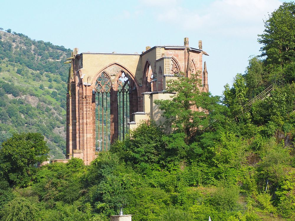 Tall gothic arches emerging from a tree-covered hill. Some of the gothic filigree is still in the upper parts of the empty windows. No roof.