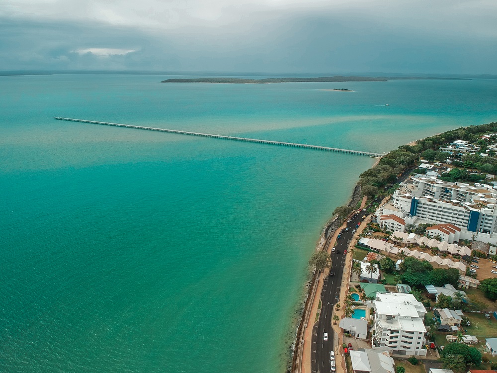 Taken from the air: Turquoise water and a curve of shore. No beach, but a road and a walking path edge the water. On the inland side of the road, many buildings of various sorts and heights. A stop on the way from Brisbane to Cairns.