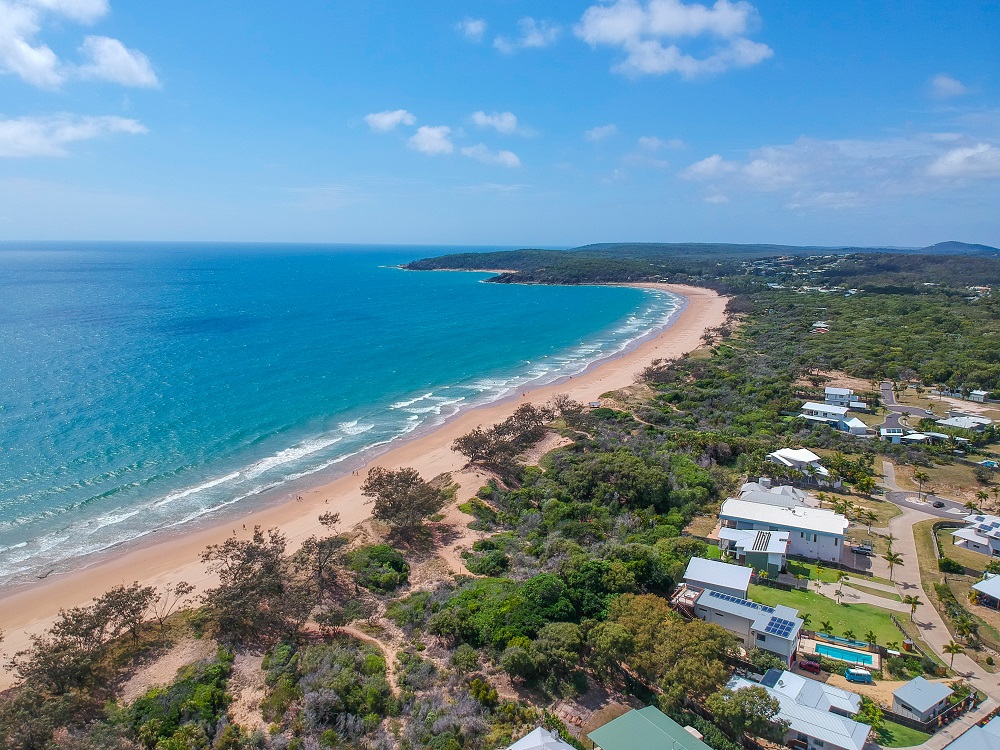 View from the sky of a long almost-straight beach lined with scrubby dunes. The land here is relatively flat. Behind the dunes are houses.