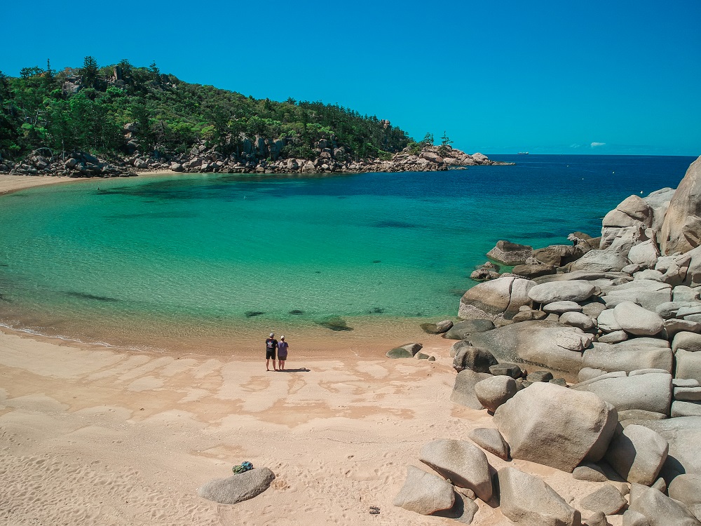 A view from the land above the beach looking down on a smooth sandy beach forming a curved bay and turquoise water, very calm. On the Brisbane to Cairns route.