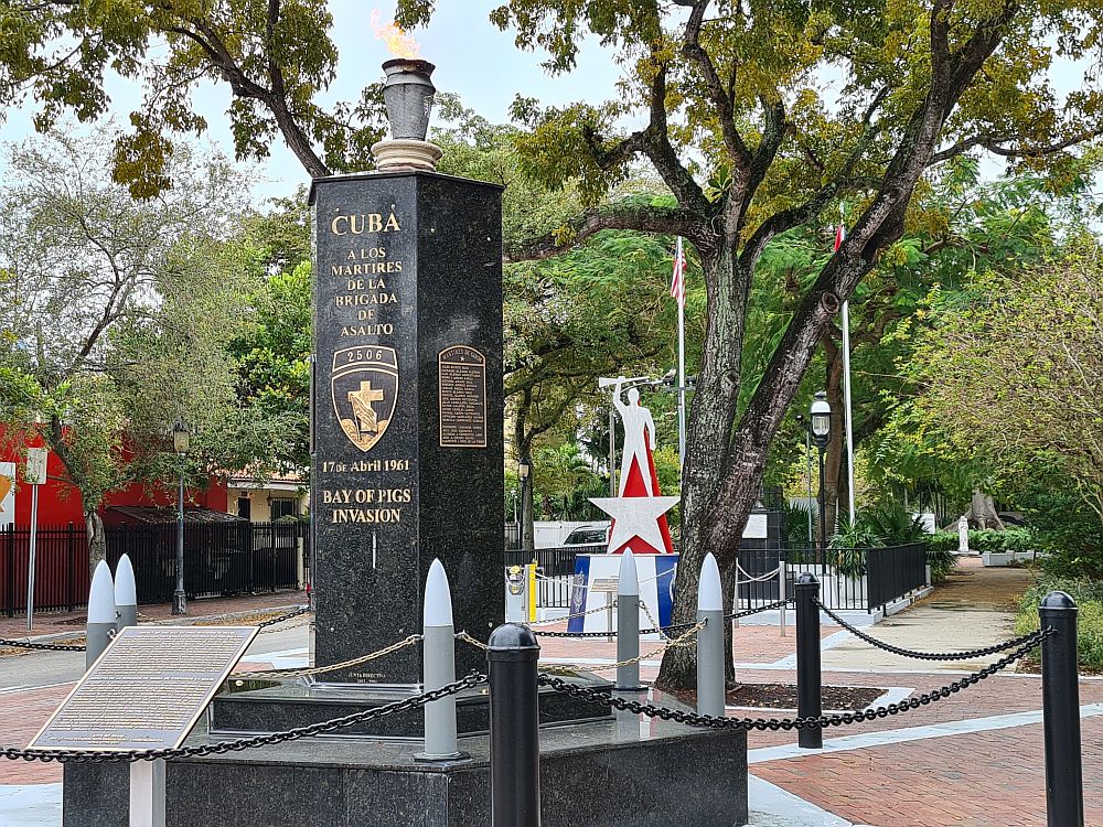 The monument in the foreground is a black column - marble, it seems - with the words "CUBA A LOS MARTIRES DE LA BRIGADA DE ASALTO" and a crest with a cross and 2506 on it. Below that "17 de Abril 1961 BAY OF PIGS INVASION". On top of the column is a torch with a flame. In the background, the other memorial shows a flat white outline of a man holding a rifle above his head. He stands on a star, his legs forming the top point of the star.