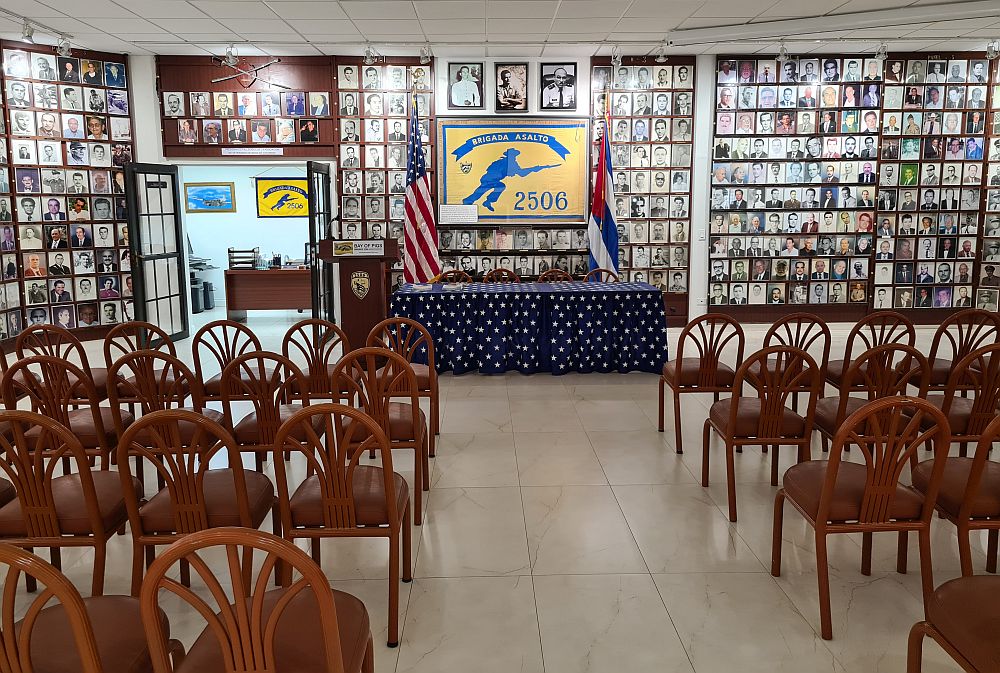 The far wall of the room is covered floor to ceiling with framed photos except for a framed flag in the center. The flag is yellow with a blue image of a running soldier holding a rifer and reads "Brigada Asalto 2506). On either side of the flag are two more flags: US and Cuba. A table in front of that is draped with the American flag. The rest of the image is filled with empty chairs facing the far wall.