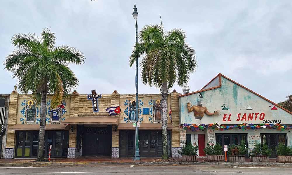 Several storefronts in Little Havana as seen from across the street. The 3 on the left seem to be a single church: images of saints above the awnings and the words "Jesus Saves" on a cross above the middle storefront. Two palm trees in front of the storefronts and also a streetlight. On the right, a storefront with a 3D image of a man's torso and the words "El Santo taqueria."