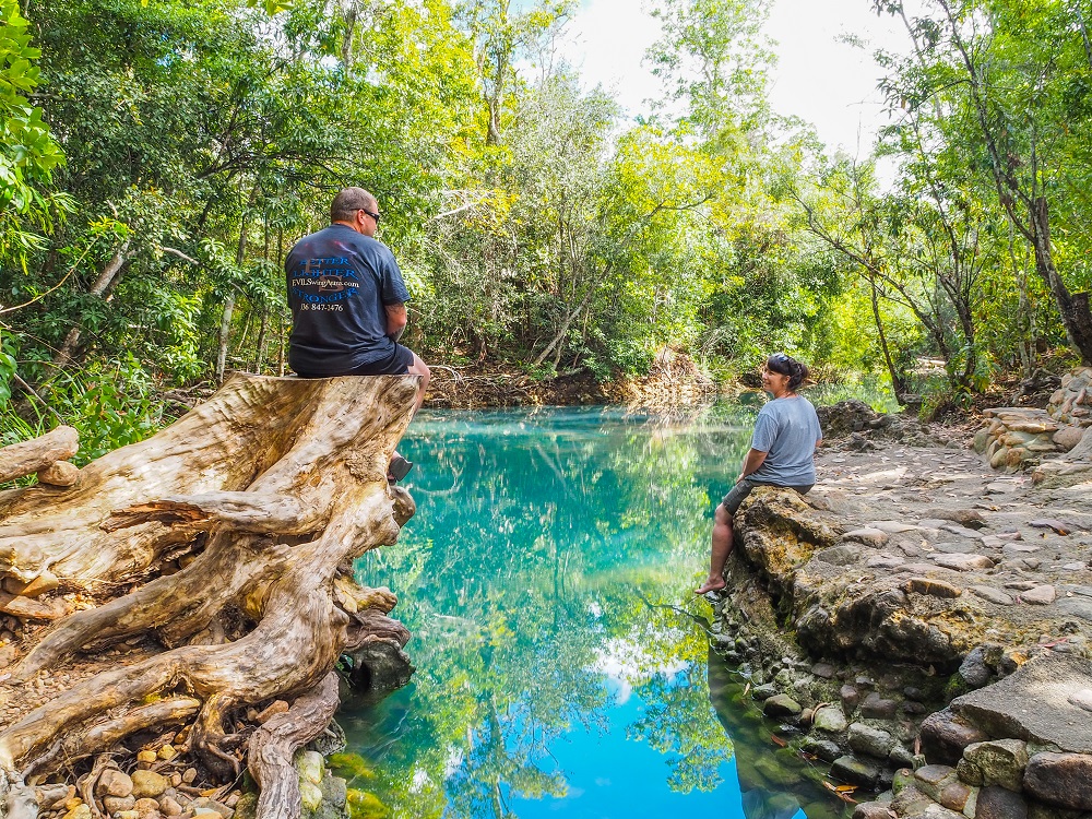 A man sits, back to camera, on a huge tree stump on the left. A woman sits on a right to the right. Between them a pool of very blue water reflects the sky and the trees surrounding the pool. 