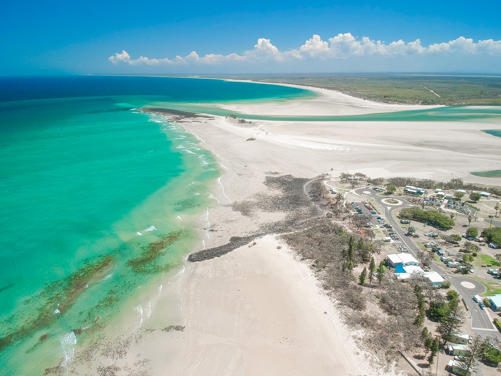 Bird's eye view of a long, deserted white sand beach, with a few buildings and campgrounds next to it. The water is a deep green-turquoise. In the distance a stream crosses the beach to the sea.