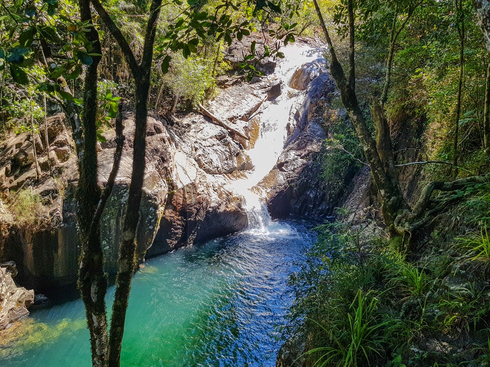 Looking down on a deep gorge filled with water and surrounded by greenery. On the opposite side, water runs in a waterfall over rocks into the gorge.