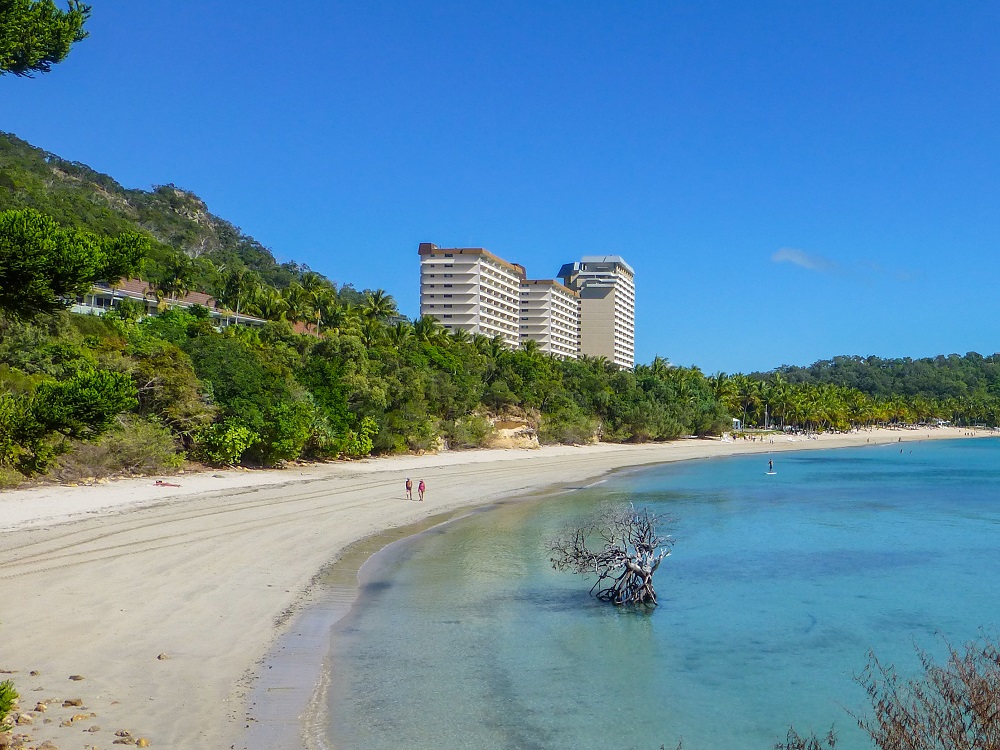 A view along a curved beach with still water. Two people walk on the beach in the distance, trees edge the beach. In the distance, behind the trees, are three tall apartment or hotel towers.