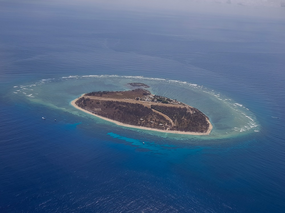 Bird's-eye view of the island, surrounded by light green water where it's shallow. The island is oblong, with a cluster of buildings on one side and a dirt airstrip down the middle.