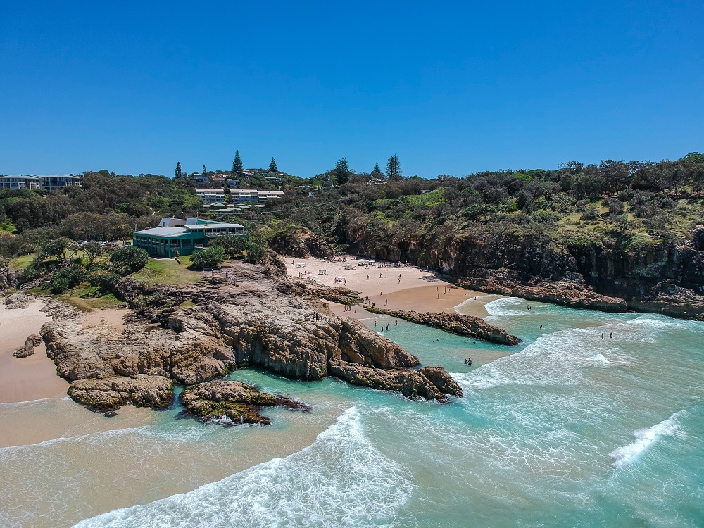 A view from the air of a cove edged by rocky points. In the cove, a sandy beach. People on the beach and also in the water.