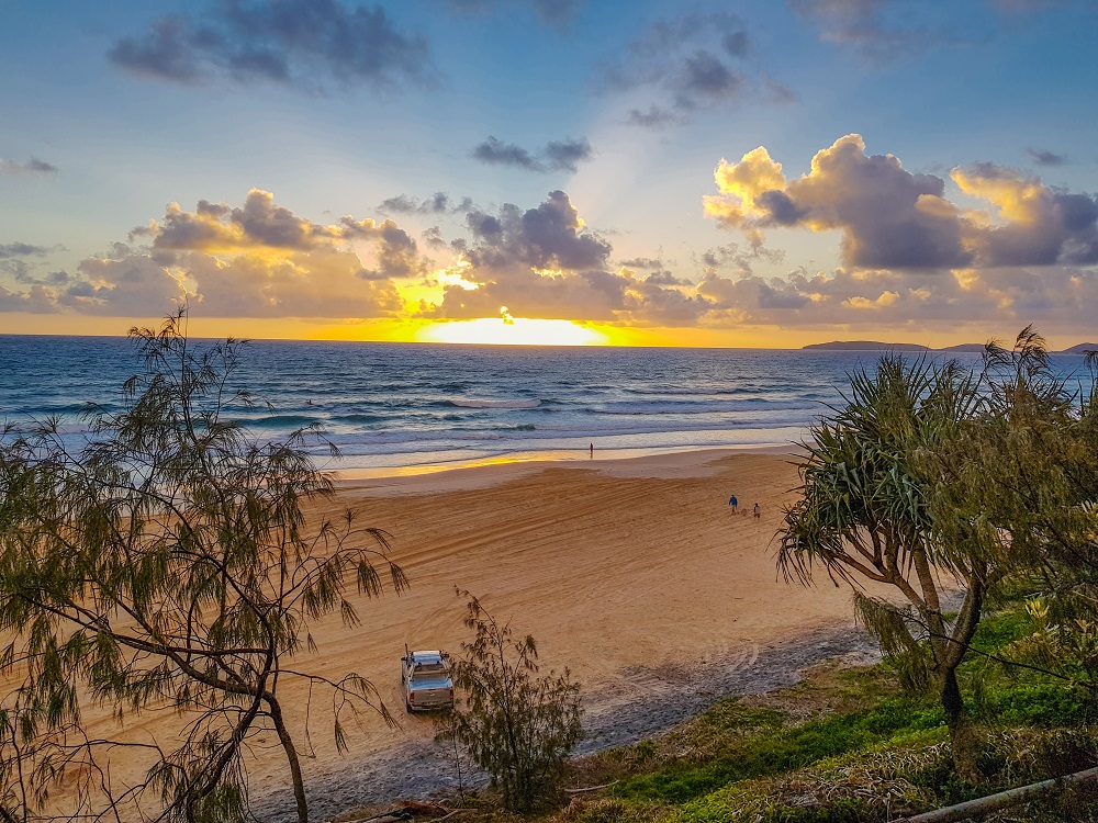 Seen from a hill behind the beach, looking over the beach to a sunset. Waves on the ocean and only a few people on the beach. 