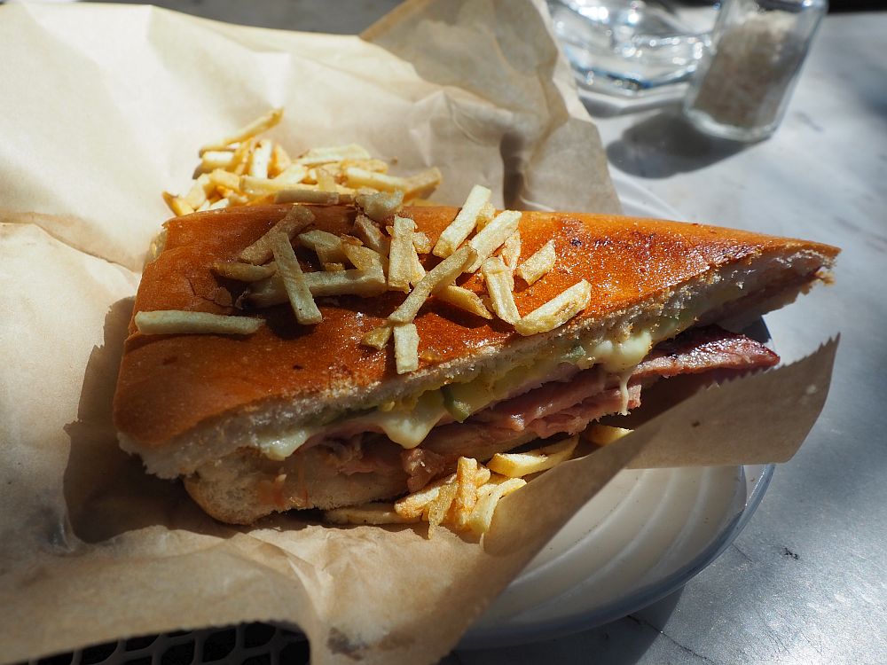 A triangular sandwich of white bread with cheese and roast ham visible on the side. Potato sticks are scattered on and around it.