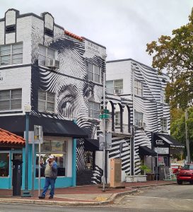 An image of a person painted on the side of a building above a storefront shop. The image is black and white and painted in curved stripes of black.