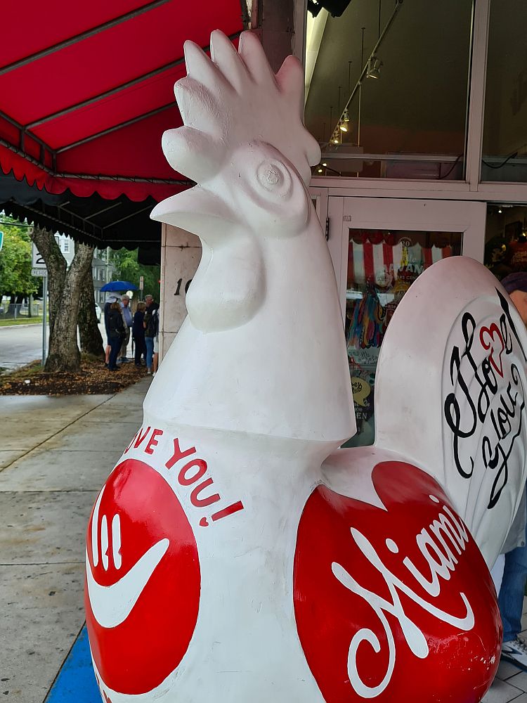 A large rooster statue, painted white. ON the chest, in red, is a circle showing the ASL sign for "I love you" and the words "I love you!" in red above the circle. On the side, the word "Miami" inside a red heart.