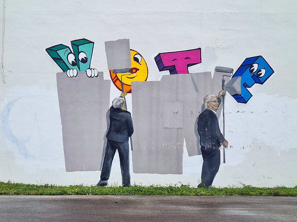 The image shows two men, life-sized, one of them recognizable as Senator Mitch McConnell. The men carry paint rollers and paint gray paint over a large colorful word "VOTE".