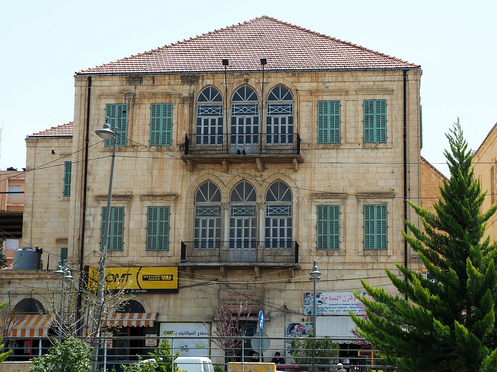 A large square structure, 3 tall stories. Shops on the ground floor, symmetrical rows of windows: 7 across, with the center 3 having arched tops and balconies. All windows are shuttered closed.