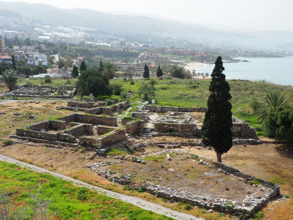 A view over several ruins, each with low walls that show where rooms in each structure were. Beyond, the coastline with many buildings on the hill between the sea and mountains behind.