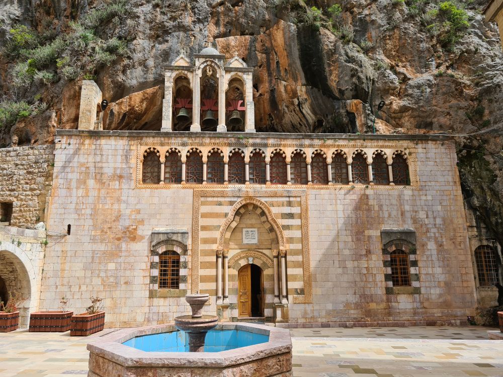 A facade that appears to lean right up against a stone cliff: white stone, arched windows, an arched entry in the center. A fountain on the courtyard in front.