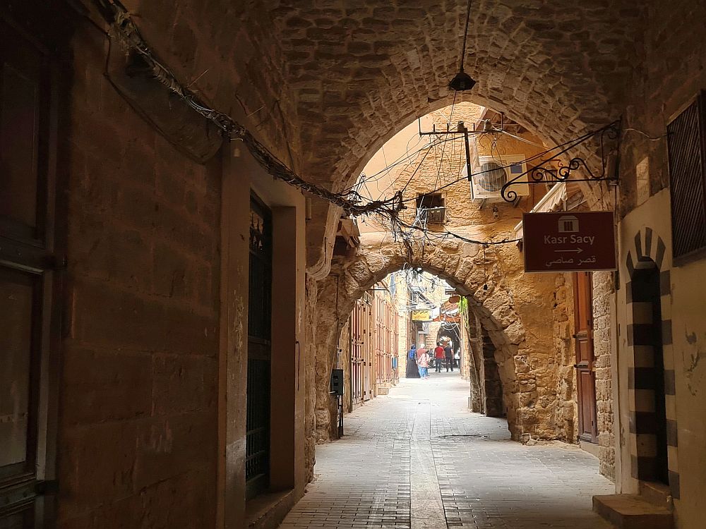 A view down a narrow road with stone walls on either side and arches in stone over it nearby. In the distance, a few people are visible in the road.
Lebanon Travel Tips