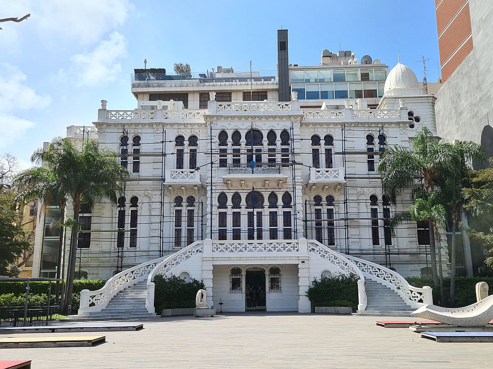 An ornate 3-story mansion with a curved stairway on either side of the entrance one story up. Arabic arches over each long window, carved banisters on the stairway and the small balconies. All painted white, with a small white dome on the right.